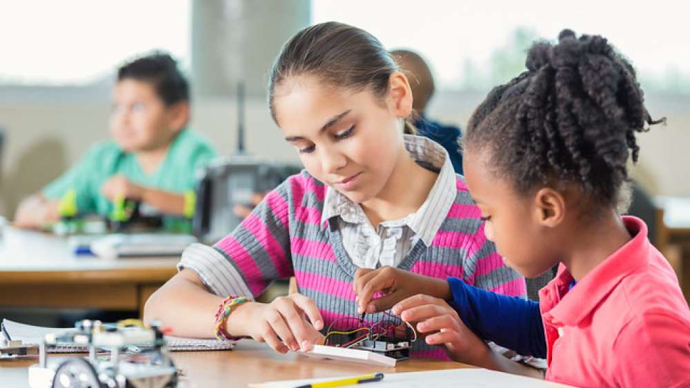 Diverse little girls in public elementary school after school science program are sitting at a table in a library. Students are using robotics kits while studying science, technology, engineering, and,  math.