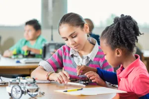 Diverse little girls in public elementary school after school science program are sitting at a table in a library. Students are using robotics kits while studying science, technology, engineering, and,  math.